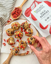 Top down image of Wildly Organic Goji Berry packaging next to goji berry protein balls with chocolate chips and oats on a parchment sheet on a round wooden cutting board with handle and next to a wood spoon and striped dishcloth.